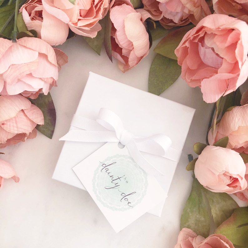 May include: A square white gift box tied with a white grosgrain ribbon bow. A square white tag hangs from the ribbon, featuring a light green circular lace pattern and the text "the dainty doe" written in a dark, elegant cursive script.