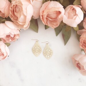 May include: A pair of gold-colored, filigree teardrop earrings with a floral design. The earrings are displayed on a white surface, surrounded by soft pink flowers and green leaves.