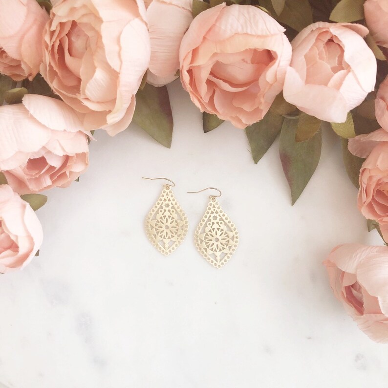 May include: A pair of gold-colored, filigree teardrop earrings with a floral design. The earrings are displayed on a white surface, surrounded by soft pink flowers and green leaves.