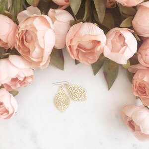 May include: A pair of gold-colored, filigree teardrop earrings with a floral design. The earrings are displayed on a white marble surface, surrounded by soft pink flowers and green leaves.