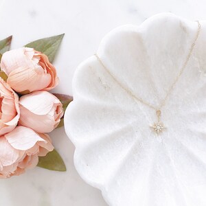 May include: A delicate gold chain necklace featuring a small, four-pointed star pendant. The star is accented with tiny, clear, sparkling stones and a central round stone. The necklace is displayed on a white, scalloped, shell-shaped dish.