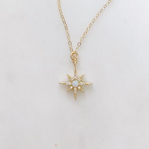 May include: A gold chain necklace featuring a starburst pendant. The pendant has a central round iridescent opal stone surrounded by small clear crystals set along the eight radiating points of the gold starburst frame.