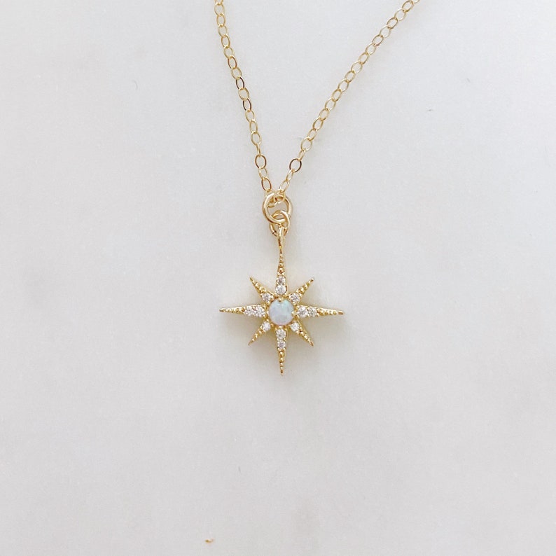 May include: A gold chain necklace featuring a starburst pendant. The pendant has a central round iridescent opal stone surrounded by small clear crystals set along the eight radiating points of the gold starburst frame.