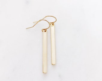 May include: A pair of gold bar earrings with a simple, minimalist design. The earrings are made of a thin, rectangular bar of metal that hangs from a small hook.