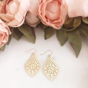 May include: A pair of gold-colored, filigree earrings with a teardrop shape. The earrings feature a floral design in the center and are set against a backdrop of pink flowers and green leaves.