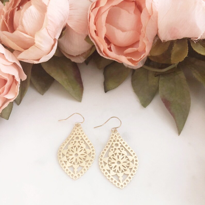 May include: A pair of gold-colored, filigree earrings with a teardrop shape. The earrings feature a floral design in the center and are set against a backdrop of pink flowers and green leaves.
