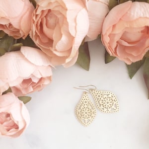 May include: Gold-colored, openwork earrings with a teardrop shape and floral design. The earrings are displayed on a white surface, with soft pink flowers in the background. The earrings have a delicate, feminine aesthetic.