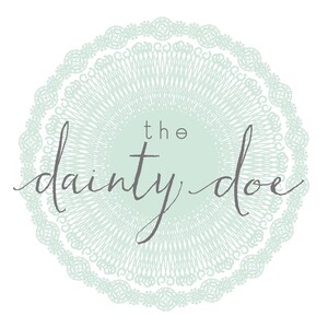 May include: A circular logo design featuring the text "the dainty doe" in a script font. The text is centered within a light green circle, surrounded by a detailed, lace-like border in a matching shade of green, all set against a white background.