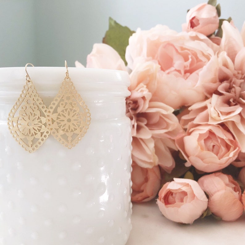 May include: Gold-colored filigree teardrop earrings with a floral design. The earrings hang in front of a white, textured jar, with a bouquet of pink roses in the background. The earrings have a delicate, openwork pattern.