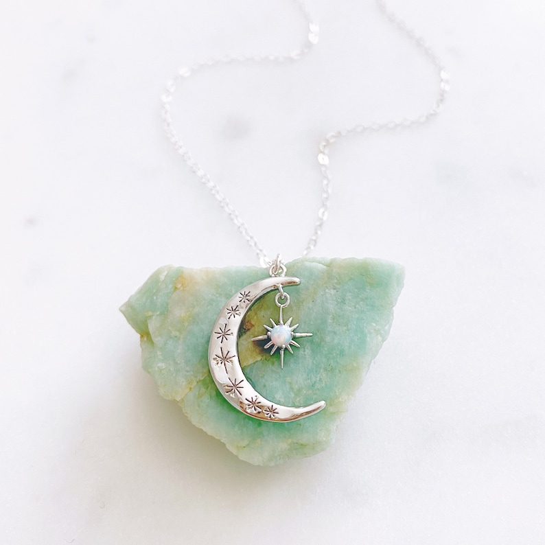 K&ouml;nnte beinhalten: Eine silberne Halskette mit einem Halbmond-Anh&auml;nger und einem Stern-Anh&auml;nger mit einem wei&szlig;en Opal-Stein. Die Halskette liegt auf einem gr&uuml;nen Steinuntergrund.