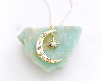 May include: A gold crescent moon necklace with a star charm featuring a white opal gemstone. The necklace is displayed on a light green rock.