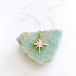 May include: A gold necklace featuring a starburst pendant with a central round opal stone and small clear accent stones. The pendant is resting against a rough, light green mineral stone, and the chain is a delicate gold link style.
