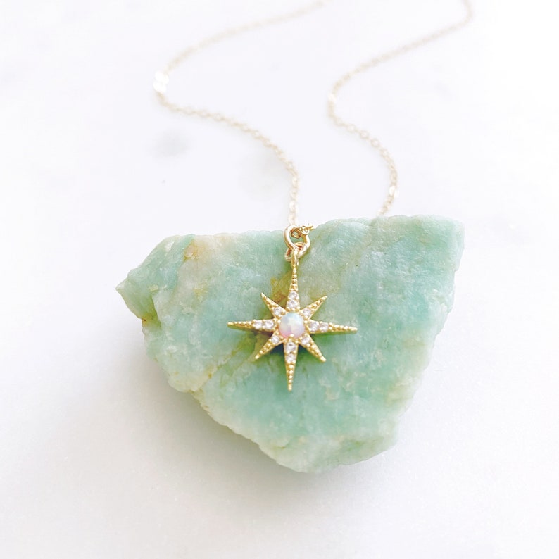 May include: A gold necklace featuring a starburst pendant with a central round opal stone and small clear accent stones. The pendant is resting against a rough, light green mineral stone, and the chain is a delicate gold link style.