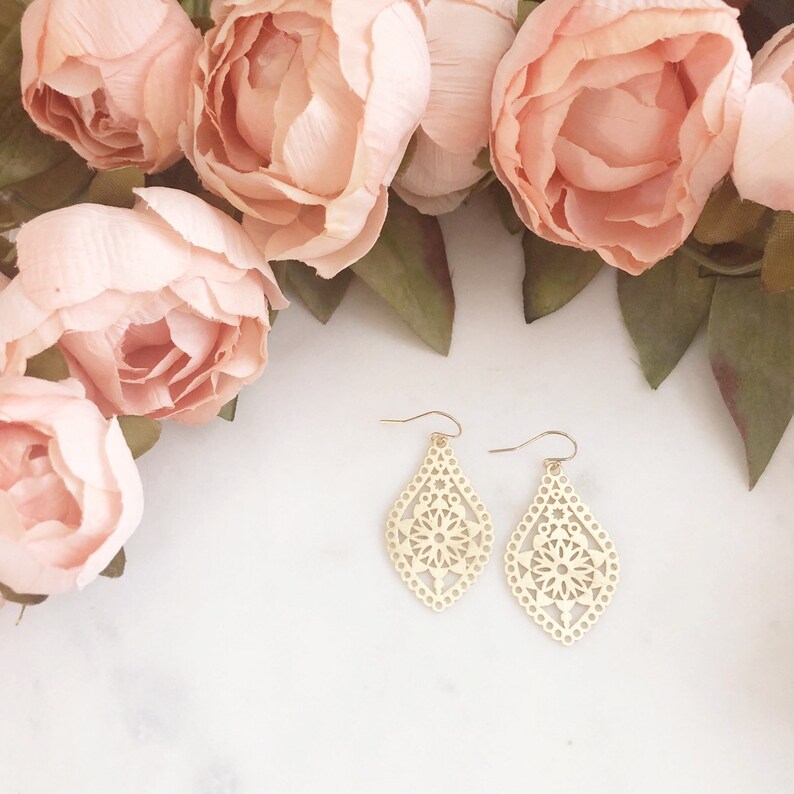 May include: A pair of gold-colored, openwork teardrop earrings with a floral design. The earrings are displayed on a white surface, with soft pink flowers and green leaves in the background.