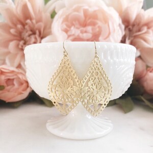 May include: A pair of gold-colored, filigree teardrop earrings with floral designs. The earrings are displayed on a white pedestal dish, with soft pink flowers in the background. The earrings have a delicate, openwork pattern.