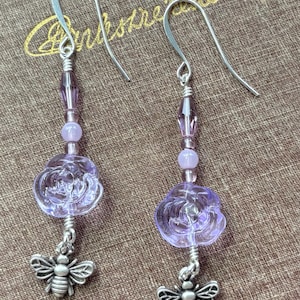 May include: A pair of dangle earrings featuring a silver bee charm, a lavender rose-shaped bead, and various purple beads. The earrings have silver hooks and are displayed on a brown surface.