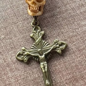 May include: A pendant featuring a carved beige skull bead positioned above a bronze-toned metal crucifix. The crucifix displays a detailed figure of a person on a cross with ornate, flared ends. The components are connected by a small metal ring.