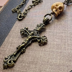 May include: A bronze-toned metal crucifix pendant featuring a detailed figure and the letters INRI at the top. The pendant is attached to a matching bronze-toned chain, which includes a small, light-colored, carved skull bead integrated into the design.
