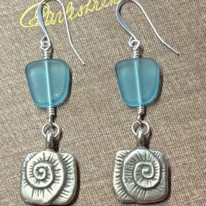May include: A pair of dangle earrings featuring light blue glass beads and silver-toned square pendants with a spiral design. The earrings have silver-colored ear wires and are displayed on a textured brown surface.