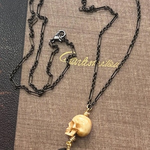 May include: A necklace featuring a dark metal chain, a cream-colored skull bead, a black faceted bead, and a round gold-toned pendant with a star design. The pendant has the text "OLD" and "FORT" embossed on its surface, connected by gold wire.