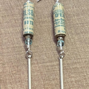 May include: Handmade dangle earrings featuring cylindrical beads made from vintage paper with text. Each earring has a silver-tone metal bar and a small blue bead. The earrings hang from silver-tone ear wires.