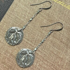 May include: A pair of silver-toned dangle earrings. Each earring features a circular pendant with a relief design and a chain of small links. The earrings hang from curved ear wires. The background is a textured, tan surface.