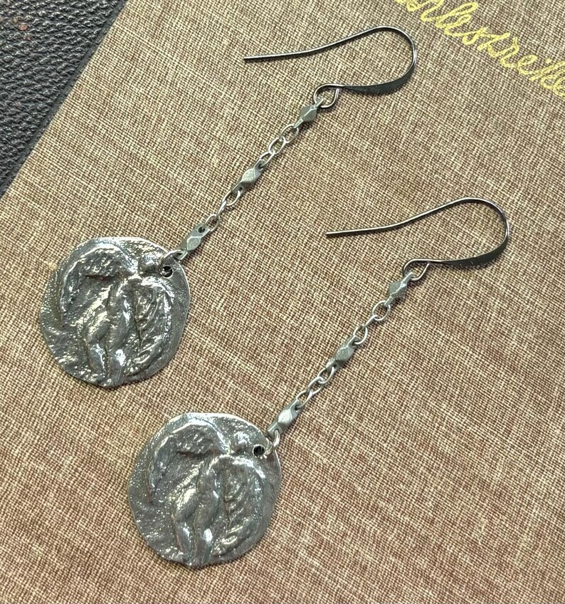 May include: A pair of silver-toned dangle earrings. Each earring features a circular pendant with a relief design and a chain of small links. The earrings hang from curved ear wires. The background is a textured, tan surface.
