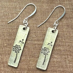 May include: A pair of silver-toned rectangular drop earrings featuring a stamped dandelion design with seeds blowing in the wind. Each earring hangs from a curved silver-toned ear wire, showcasing a textured metallic finish on the rectangular bar.