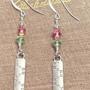 May include: A pair of dangle earrings with silver-tone hooks. Each earring features a strand of pink, green, and clear beads, leading to a rectangular, silver-tone charm with a floral pattern. The earrings are displayed on a textured brown surface.