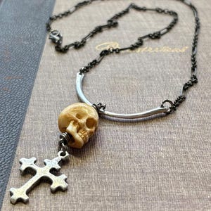 May include: A necklace featuring a dark metal chain connected to a silver-toned curved bar. A beige skull bead is suspended from the bar, with a silver-toned cross charm hanging from the bottom of the skull. The chain has a lobster claw clasp.