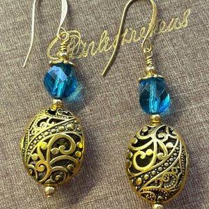 May include: A pair of gold-colored earrings with intricate filigree oval pendants and faceted blue beads. The earrings have gold-colored ear wires and small gold beads. The background is a neutral brown.