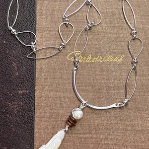 May include: Silver-toned necklace with a fish pendant. The necklace features a series of oval and teardrop-shaped links. The pendant is a white fish with a pearl and brown bead accent. The necklace is displayed on a brown surface.