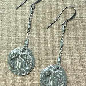 May include: A pair of silver-toned dangle earrings. Each earring features a circular pendant with a relief design of figures and foliage, suspended from a chain and a hook. The earrings are displayed against a textured, neutral background.