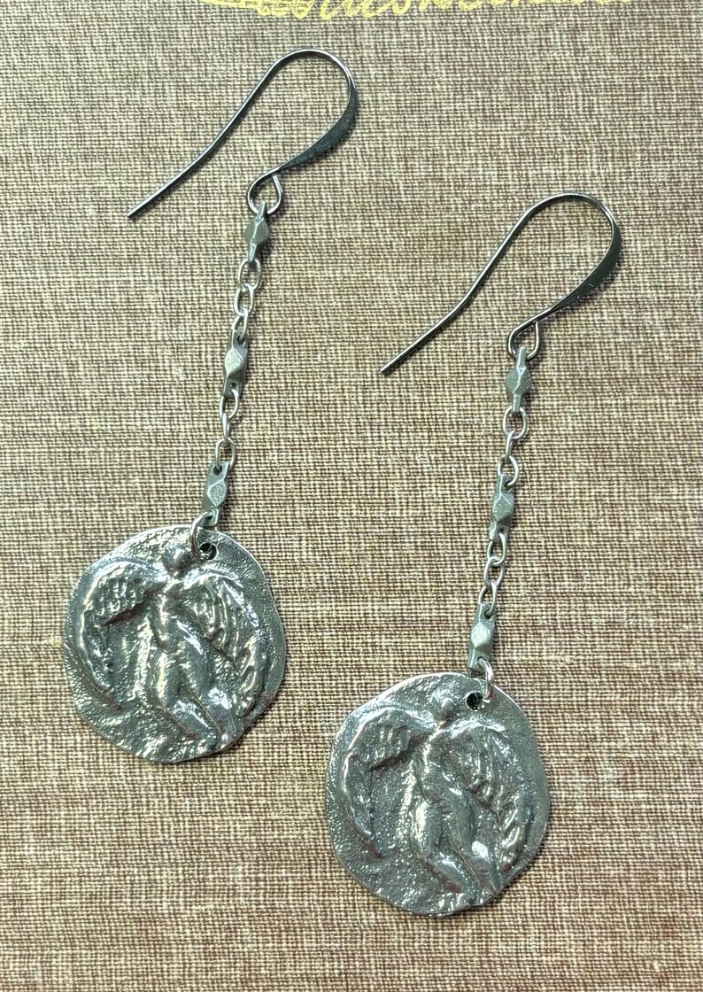 May include: A pair of silver-toned dangle earrings. Each earring features a circular pendant with a relief design of figures and foliage, suspended from a chain and a hook. The earrings are displayed against a textured, neutral background.
