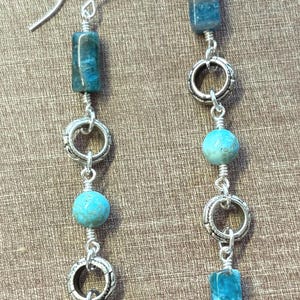 May include: A pair of dangle earrings featuring turquoise and silver-tone metal. Each earring has a silver hook, a turquoise-colored rectangular bead, a silver ring, a turquoise sphere, another silver ring, and a turquoise rectangular bead.