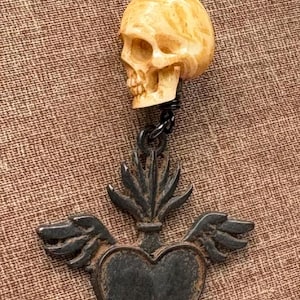 May include: A pendant necklace featuring a carved beige skull bead suspended above a dark metallic heart charm. The heart charm is adorned with small wings on either side and a flame-like crest at the top, all attached to a dark chain link.