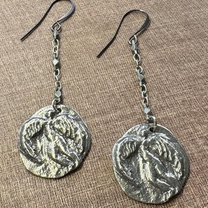 May include: A pair of silver-toned dangle earrings. Each earring features a textured, circular pendant with a relief design, suspended from a chain and a hook. The earrings have a vintage or antique aesthetic.