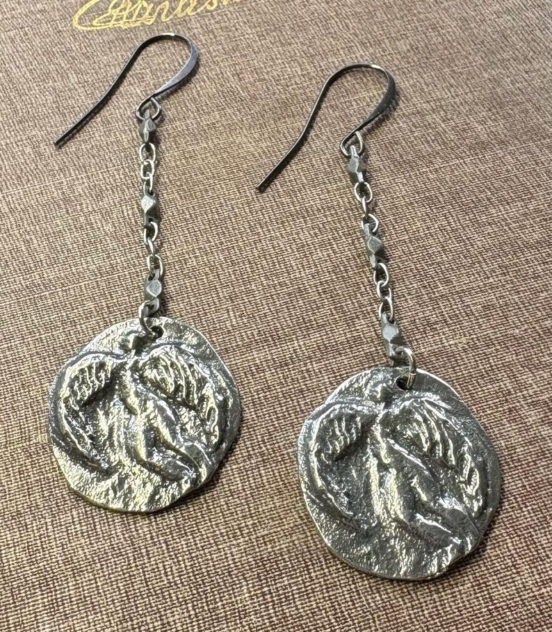 May include: A pair of silver-toned dangle earrings. Each earring features a textured, circular pendant with a relief design, suspended from a chain and a hook. The earrings have a vintage or antique aesthetic.