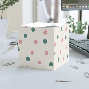 May include: A square memo cube featuring a cream-colored background with a repeating pattern of small, hand-drawn style dots in muted pink and dark teal. The paper block has a slightly curled top sheet, showcasing a stack of individual note pages.
