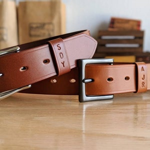 May include: Two brown leather belts with silver buckles. The belt on top has the name "SDY" embossed on it. The belt on the bottom has the name "AJR" embossed on it.