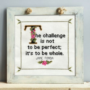 May include: Cross-stitch artwork in a distressed white frame. The text reads "The challenge is not to be perfect; it's to be whole." A floral "T" and a pink flower accent the quote by Jane Fonda. Two rope knots adorn the top.