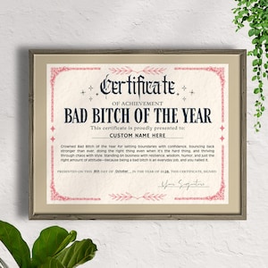 May include: Framed certificate with the text "BAD BITCH OF THE YEAR" in navy blue. The certificate has a pink decorative border and includes space for a custom name. The certificate is presented on the 8th day of October, in the year of 2026.