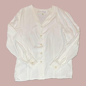 May include: A white long-sleeve blouse with a V-neck and button-down front. The blouse has a relaxed fit and features buttoned cuffs. The fabric appears to be a lightweight material. The blouse is a classic style.