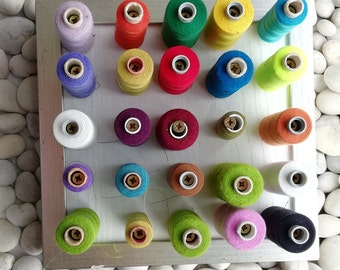 May include: A silver frame with a white background holds 24 spools of thread in various colours. The spools are arranged in a grid pattern.