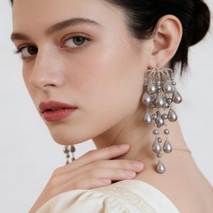 Puede incluir: Pendientes colgantes plateados con cuentas grises en forma de lágrima. Los pendientes tienen un diseño delicado y adornado con múltiples hilos de cuentas que caen en cascada. Los pendientes se llevan en la oreja de una persona.