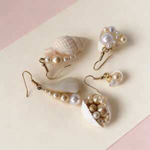 May include: Five pairs of handcrafted earrings featuring seashells and faux pearls. The earrings have gold-colored hooks and vary in design, with some incorporating small, gold-colored beads. The color palette includes white, cream, and gold.