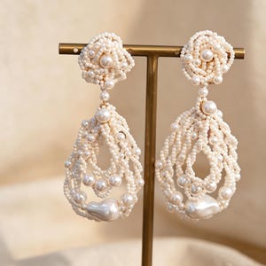 Puede incluir: Un par de elegantes pendientes de cuentas de color blanco roto. Cada pendiente presenta una parte superior en forma de flor, una delicada cadena y un diseño en forma de lágrima. Los pendientes están adornados con numerosas cuentas pequeñas y perlas más grandes y brillantes, creando un accesorio sofisticado y llamativo.