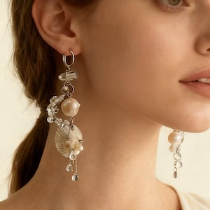 Puede incluir: Pendientes largos y colgantes con un tema de conchas marinas y perlas. Los pendientes presentan un aro plateado, cristales transparentes, una gran perla color melocotón y una pequeña concha. Cuentas y cristales transparentes adicionales cuelgan debajo.