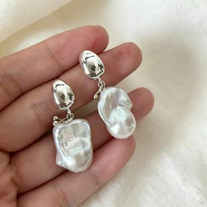 Op de afbeelding: Zilverkleurige oorhangers met barokparels. De oorbellen hebben een gepolijst, metalen bovenstuk dat door een kleine ketting met de parel is verbonden. De parels zijn onregelmatig van vorm en hebben een crèmekleur.