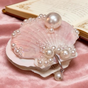 May include: A pink seashell adorned with numerous pearls of varying sizes. A large pearl sits atop the shell, with smaller pearls outlining the edges. A silver-toned pendant with a dangling pearl is attached.